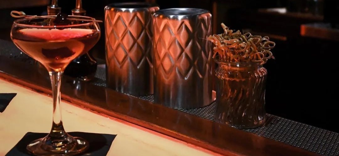 A craft cocktail on the bar at Lucy's Lounge, beside copper shaker tins